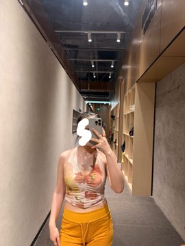 A mirror selfie of a person in a yellow top and orange pants standing in a modern hallway, with their face obscured by a white graphic.