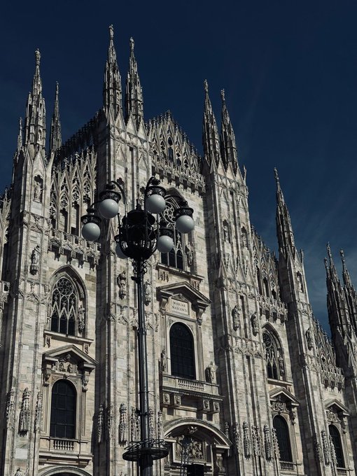 A Gothic-style building with a pointed roof and tall spires stands against a clear blue sky, accompanied by a street lamp on the left side.