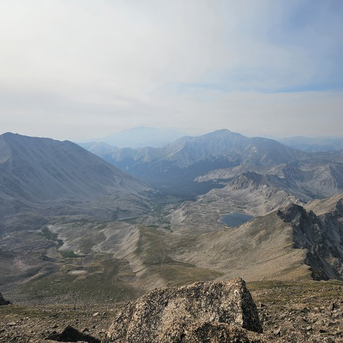 A panoramic view from a high mountain summit showing rocky alpine terrain, a valley with lakes, and distant mountain ranges under a partly cloudy sky.
