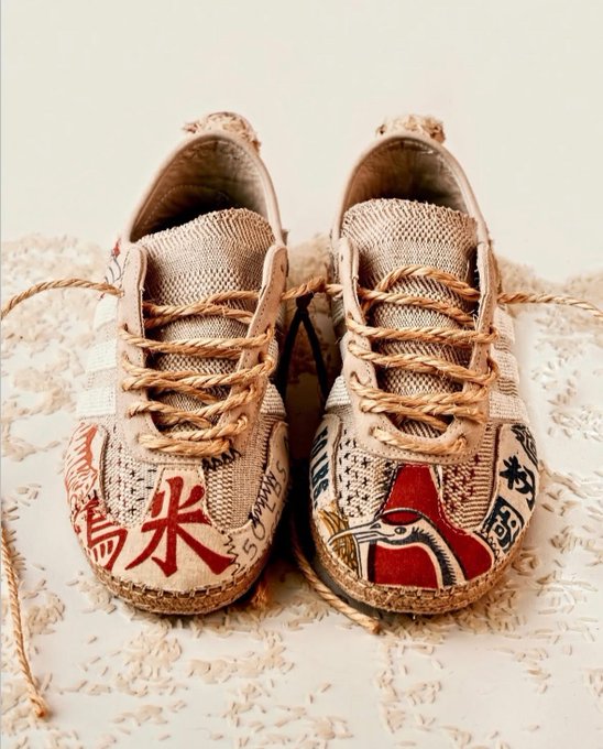 A pair of artistic fabric sneakers displayed on rice grains, featuring beige canvas uppers, braided rope laces, and colorful printed designs including a red bird and Asian characters.
