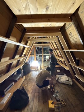 Two people are working inside a wooden attic or loft space, illuminated by a bright light, with tools, bags, and equipment scattered on the floor.