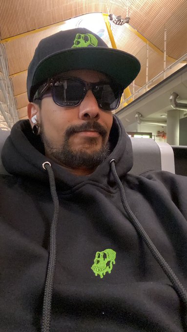 A man wearing a black hoodie with a green skull logo and matching cap takes a selfie, rocking sunglasses and AirPods in what appears to be an airport terminal. The "Gn fam" caption matches the casual, laid-back vibe of his comfortable travel attire and relaxed pose.
