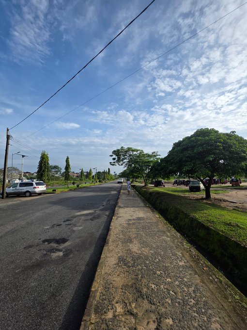A sunny outdoor street scene showing an asphalt road with potholes, a concrete drainage divider, parked cars, and lush green trees under a blue sky with scattered clouds.