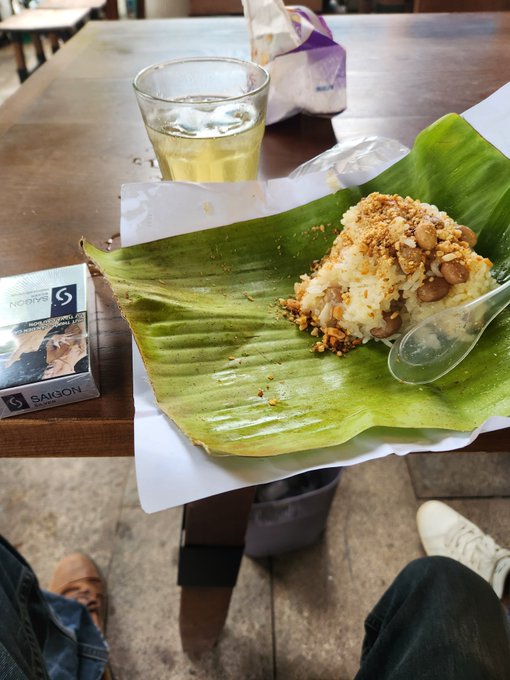 A mound of rice and beans topped with crushed crunchy bits sits on a banana leaf with a plastic spoon, while a drink and a “SAIGON” travel brochure lie on the table.