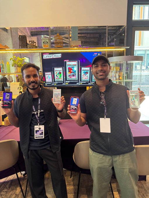 Two men wearing vests and lanyards smile and hold up trading cards in front of a digital screen displaying 'LATEST PULLS'.