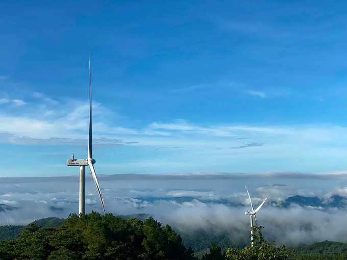 A scenic landscape of a wind turbine and a second smaller turbine on a mountain ridge, with a sea of clouds below and a bright blue sky above.