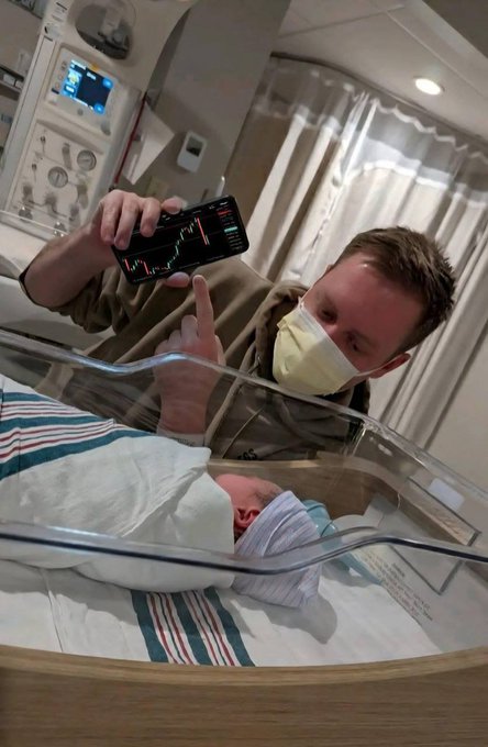 A heartwarming photo of a father wearing a face mask pointing at a stock/crypto chart on his smartphone, showing it to his newborn baby in a hospital bassinet.