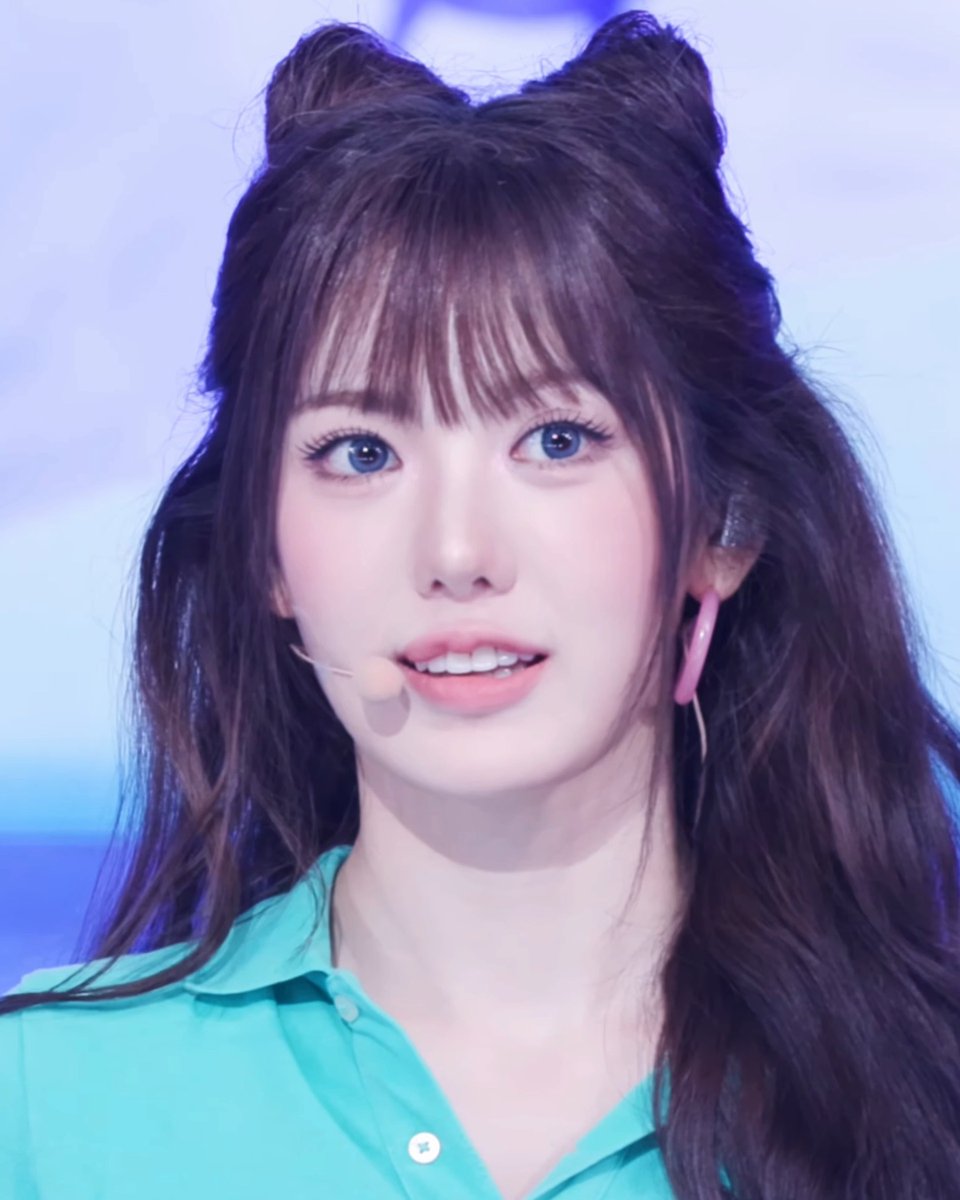 A young woman with long dark hair styled with two buns on top, bangs, and blue eyes, wearing a teal collared shirt and a headset microphone, appears to be performing or speaking on stage.