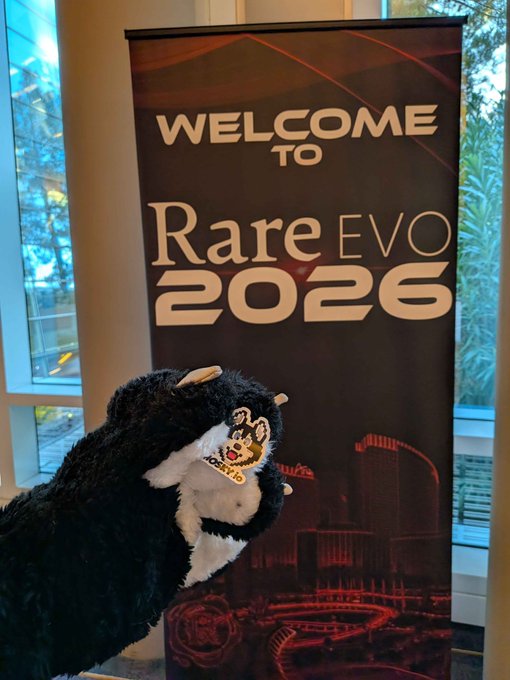 A plush mascot stands in front of a banner that reads 'WELCOME TO Rare EVO 2026', featuring a city skyline graphic.