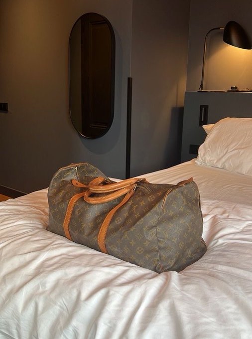 A worn Louis Vuitton monogram Speedy duffel bag resting on white hotel bedding, with a dark oval mirror and a wall-mounted reading lamp visible in the background.
