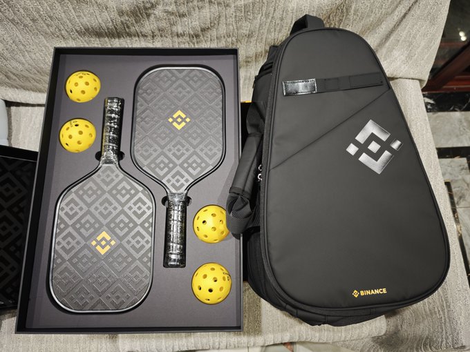 A Binance-branded pickleball set displayed in a box with two paddles and four yellow balls, alongside a black carrying bag with the Binance logo.
