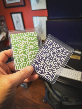 A hand holding two plastic-sleeved playing cards featuring abstract maze-like patterns, one in green and one in dark blue.