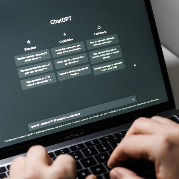 A photograph showing hands typing on a MacBook Air laptop, with the screen displaying the ChatGPT interface. The interface features columns for 'Examples' and 'Capabilities', a 'Limitations' column, and a query prompt asking 'How do I make an HTTP request in Javascript?'.