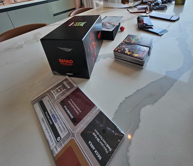 A photograph of a tabletop showing the 'Shaq' trading card game, including a black 'SHAQ' box, stacks of cards, and player aid sheets.