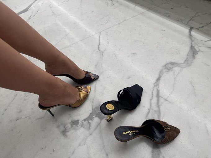 A person's legs in Saint Laurent high-heeled slingback shoes, with additional black and brown YSL shoes displayed on a marble floor.