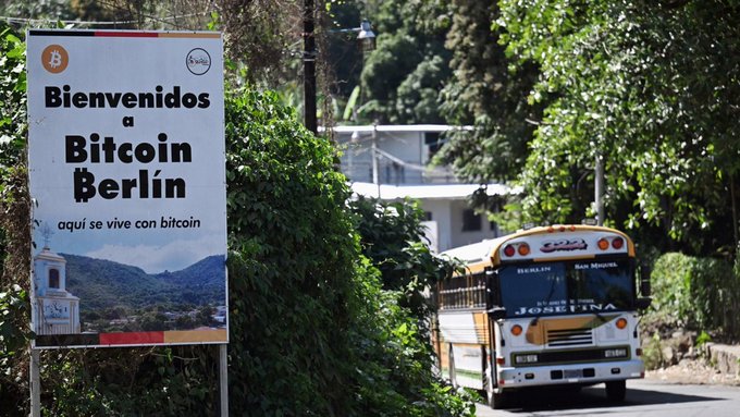 A roadside sign reads "Bienvenidos a Bitcoin Berlín - aquí se vive con bitcoin" welcoming visitors to El Zonte, the Bitcoin Beach town in El Salvador, with a yellow "JOAQUIN" school bus driving past on a lush green road.
