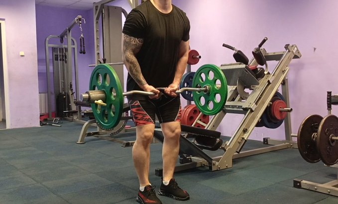 A man in a gym performing a barbell curl with an EZ-curl bar loaded with green weight plates, surrounded by various weightlifting equipment.