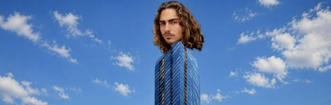 A young man with long wavy brown hair stands in a blue pinstripe suit against a vivid blue sky filled with scattered white clouds, embodying the confident "Top Floor Boss" persona of the $TJR Solana project.