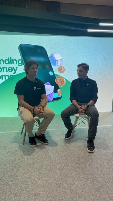 Two men seated on stools in front of a large green display featuring a smartphone and various digital icons, appearing to be at a tech or business presentation event.