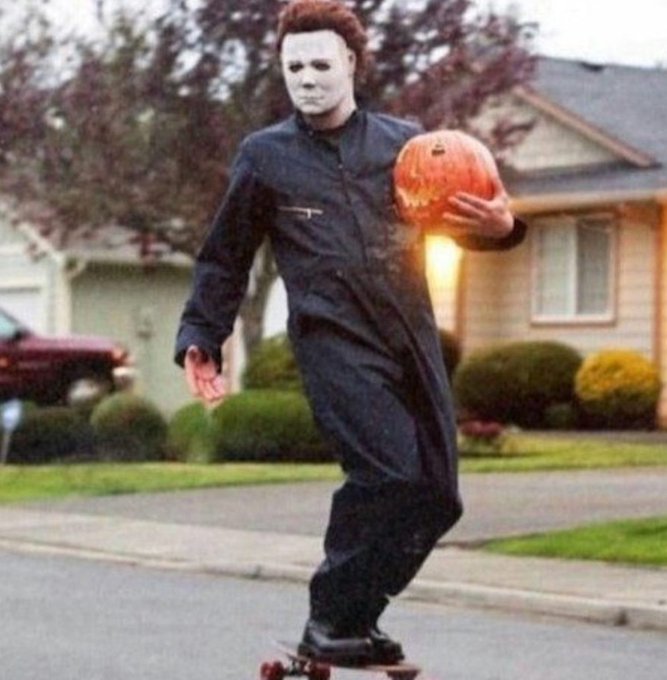 A person dressed as Michael Myers from Halloween rides a skateboard while holding a carved jack-o'-lantern down a suburban street.