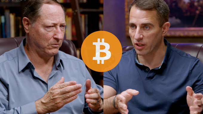 Two men in blue shirts sit side-by-side with a Bitcoin logo between them, appearing to discuss cryptocurrency.