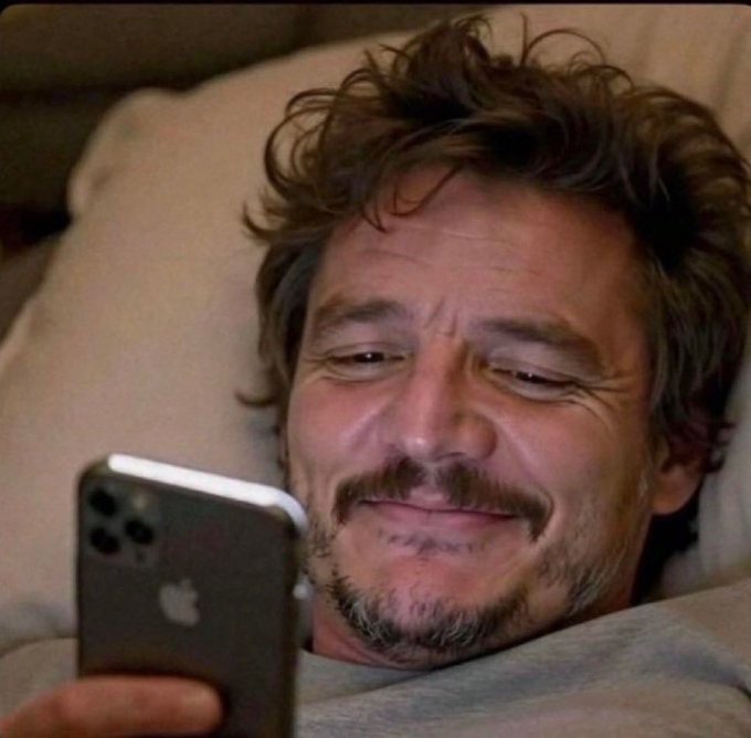 A close-up selfie of actor Pedro Pascal lying down and smiling while looking at an Apple iPhone.