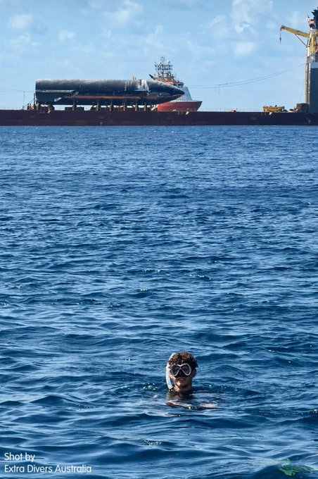 A snorkeler wearing a mask floats in deep blue water with a diving mask visible above the surface, while in the background a large transport ship labeled "Ship40" is docked at a port with industrial cranes nearby. The caption "Shot by Extra Divers Australia" appears in the bottom left corner, crediting the underwater exploration company capturing this unique juxtaposition of recreational diving and maritime industry.
