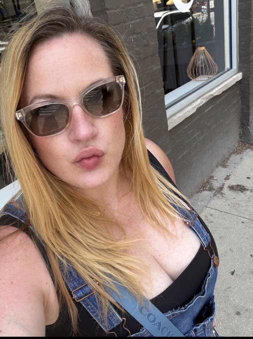 A blonde woman wearing sunglasses and a denim overall dress over a black top takes a selfie on a sidewalk in front of a dark brick building.