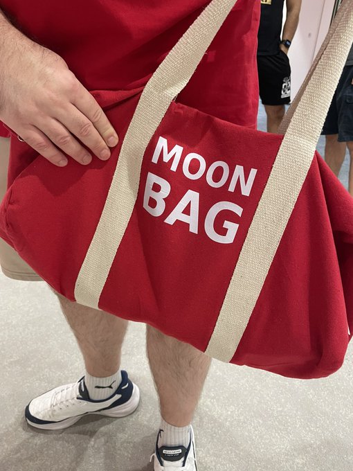 A person carrying a large red canvas tote bag with the white text 'MOON BAG' printed on it, wearing white Nike sneakers.