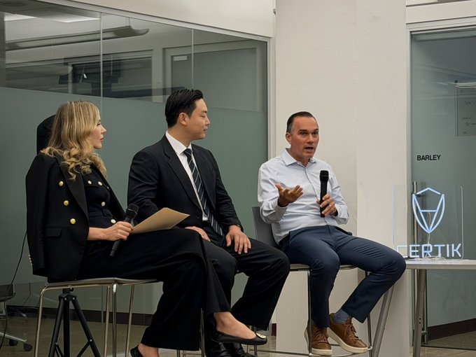 Three people sitting on stools during a panel discussion; a man on the right speaks into a microphone, with a CERTIK logo visible on a stand beside him.