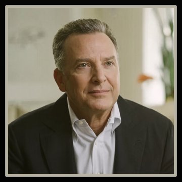 A middle-aged man with short, graying hair and a serious expression, wearing a black blazer over a white collared shirt, sits in a softly lit interior space, likely representing Steve Witkoff as mentioned in the accompanying post about his reported $107 million in 2025 income from his World Liberty Financial holding company stake.