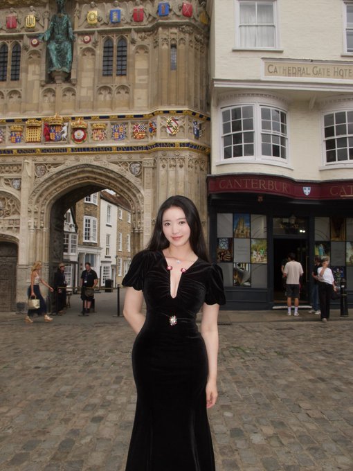 A young woman in a long black dress stands in front of Canterbury Cathedral Gate and the Cathedral Gate Hotel, with a shop labeled "CANTERBURY" visible behind her.