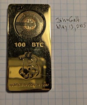 A gold-colored bar engraved with '100 BTC' and a Bitcoin logo, photographed on grid paper with a handwritten note reading 'JohnGalt May 13, 2025'.