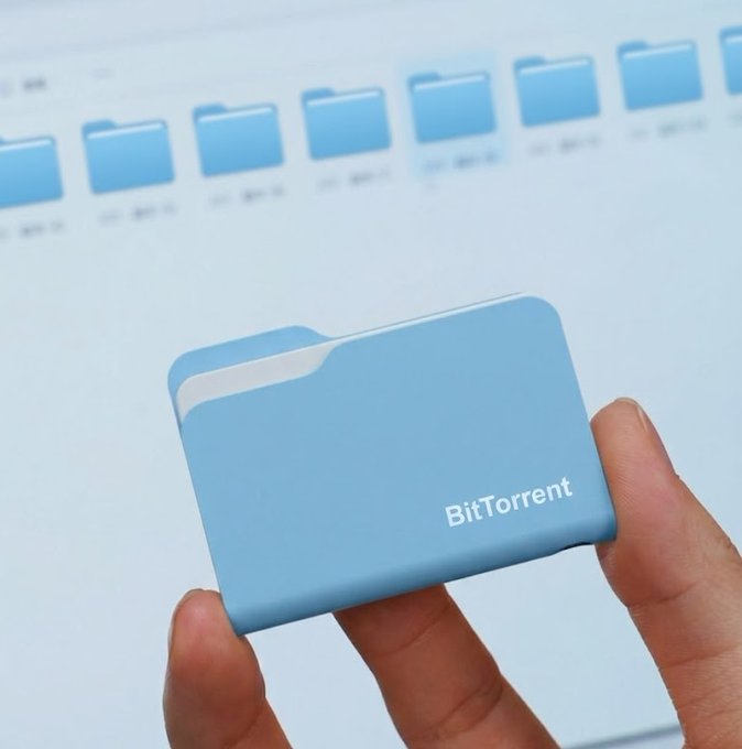 A hand holds a light blue physical folder labeled "BitTorrent" in front of a computer screen displaying multiple folder icons, visually representing the file storage and organization capabilities that underpin BitTorrent Token's decentralized utility for faster downloads and dApp integration.