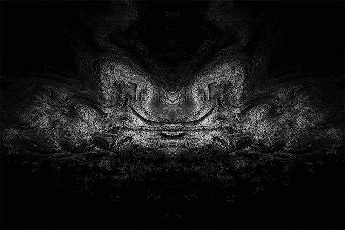 A dark, abstract symmetrical image resembling mirrored smoke, clouds, or ink swirling in black and grayscale tones.
