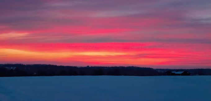 A scenic landscape photograph of a vibrant pink and orange sunset over a snow-covered field with a silhouetted treeline in the distance.