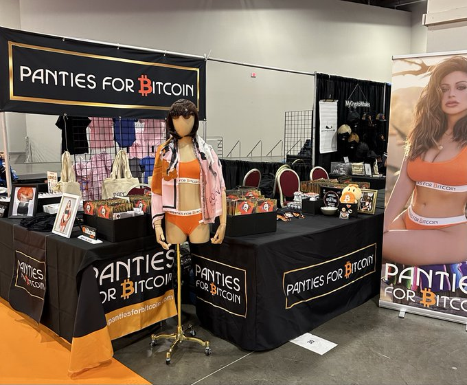 A trade show booth for 'PANTIES FOR BITCOIN' features a mannequin wearing an orange bikini and denim jacket, displaying branded merchandise and tote bags on tables with black tablecloths.