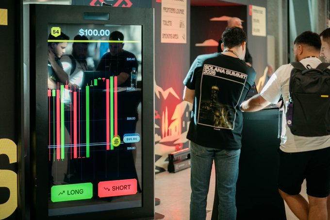 At a bustling Solana Summit 2024 event, attendees gather around a large interactive display showing a trading interface with candlestick charts and clear "LONG" and "SHORT" buttons, directly illustrating the post's question of "long, or short?" The screen prominently displays "$100.00" and shows price levels like $96.40, $95.80, $96.60, and "S4" in the top corner, while a person wears a black "SOLANA SUMMIT 2024" t-shirt featuring the Burj Khalifa design, visually tying together the promotion of Solflare Perps powered by PhoenixTrade for trading 50+ markets directly within the Solflare wallet.