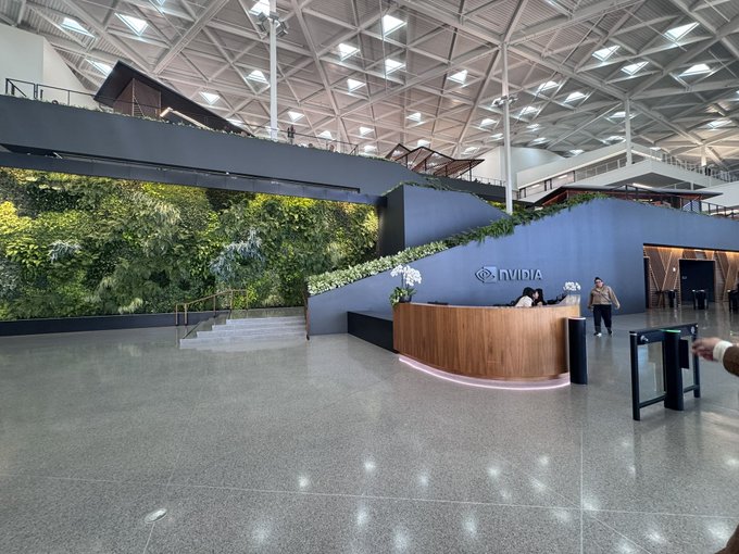 A spacious modern corporate lobby featuring a lush vertical green wall, a wooden reception desk with the 'NVIDIA' logo, security gates, and a geometric skylit ceiling.