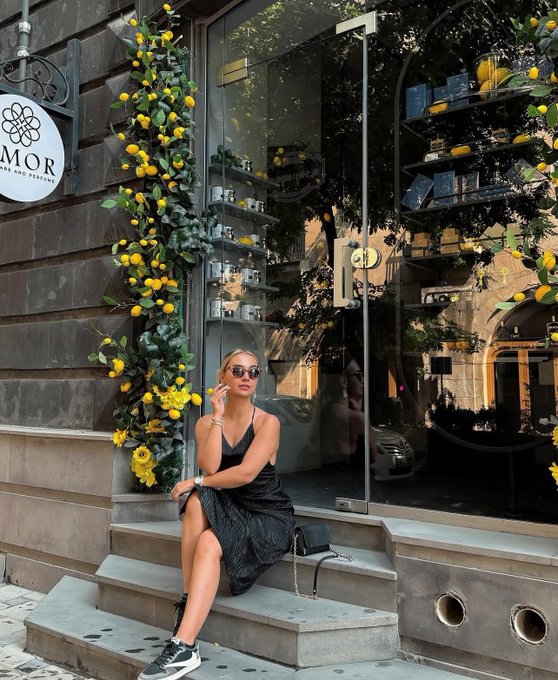A woman in a black dress, sunglasses, and sneakers sits on stone steps outside a boutique named 'MOR', framed by greenery with yellow lemons.