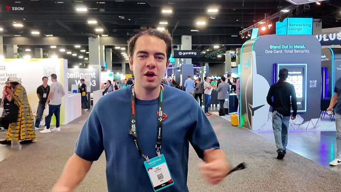 A man wearing a TRON-branded lanyard speaks on camera at a busy convention hall, with visible signage including "TRON," "Networking Lounge," "Stand Out in Metal, One Card. Total Security," and booth numbers like "56" and "37" in the background. The setting appears to be a tech or blockchain industry conference, aligning with the post about TRON DAO's efforts to expand cross-border e-commerce partnerships by integrating native TRON gateways for online merchants.