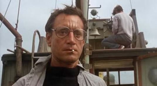 A man with glasses and a cigarette in his mouth stands on a wooden boat structure with another person working in the background.