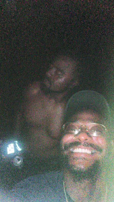 A grainy, low-light selfie of two men smiling, one in the foreground wearing glasses and a cap, with a phone screen visible in the background.