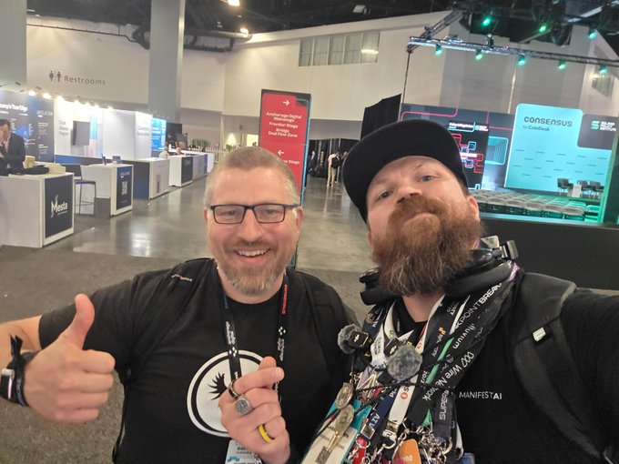 Two bearded men pose for a selfie at a convention; the man on the left wears glasses and gives a thumbs-up, while the man on the right wears a black cap and has headphones around his neck, with event booths and a 'consensus' by CoinDesk sign visible in the background.