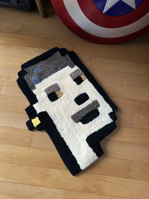 A pixel art rug resembling a smartphone with a minimalist face design lies on a wooden floor, with a Captain America shield visible in the background.
