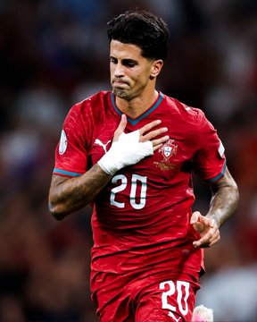 A soccer player wearing a red Serbia national team jersey with the number 20 celebrates on the field with his hand over his heart, with tattoos visible on his arms.