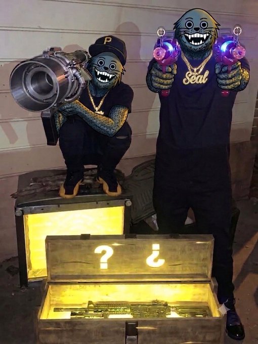 Two people posing with cartoon monkey/ape masks over their faces, one holding a large camera lens and the other holding glowing purple lights, standing next to an open treasure chest containing gold items.