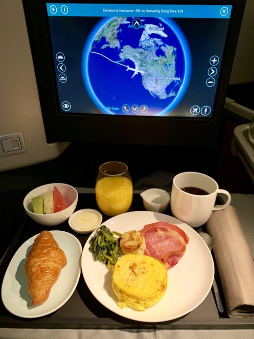 An in-flight meal served on an airplane tray featuring a croissant, fruit, an omelet with ham and vegetables, juice, and coffee, with a flight map showing a plane traveling from Anchorage toward Vancouver.