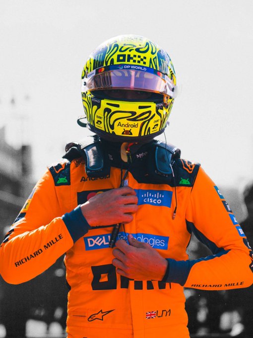 A McLaren F1 driver adjusts his race suit and HANS device, wearing a vibrant helmet with "Android" branding, while the orange racing suit prominently displays sponsors like Cisco, Dell Technologies, Richard Mille, and the number 4, signaling his preparation for the Bahrain Grand Prix.