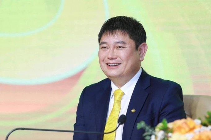 A smiling man in a dark blue suit and bright yellow tie speaks at a podium during what appears to be a press event or corporate announcement, with a small pin visible on his lapel.
